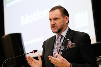 Nick wears dark suit with colourful patterned tie as he presents with hands raised at the lectern.