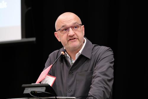 Anthony Bellanger wears  a dark long sleeved shirt and dark glasses as he speaks into mic at lectern. 