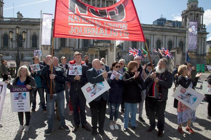 A group of people clapping while standing in front of a red 'NUJ' banner on a sunny day.