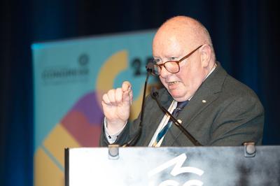 Seamus Dooley wears dark glasses and suit as he speaks into mic at lectern.