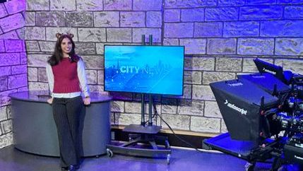 Hana Williams stands beside a television and desk in a studio with blue lighting.
