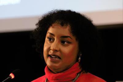 Huda wears a bright red top as she addresses delegates at the lectern