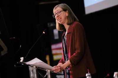 Laura wears a dark red top and spectacles as she speaks at lectern. 