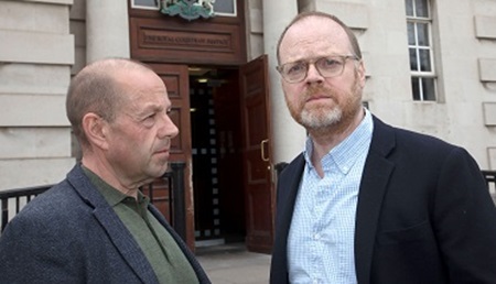 Barry wears a dark coloured shirt and suit jacket whilst looking at Trevor. Trevor wears a light blue shirt and dark coloured suit jacket, dark glasses as both are photographed.