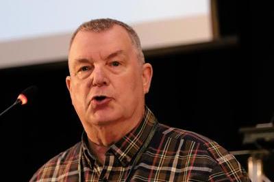 David wears a dark checked shirt as he speaks at lectern mic