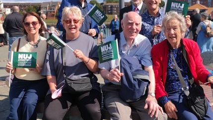 Members of the Sunderland, Shields and Hartlepool branch pose outside the river Wear with green NUJ flags with cloudy blue skies in the background.