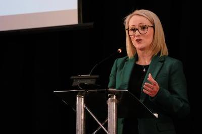 Rebecca presents from lectern, she wears a green blazer and dark glasses. 