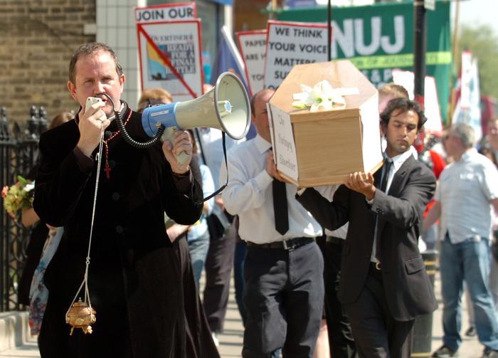 Jonathan Lovett dressed as a priest holding a microphone while suited men carry a coffin in the background.