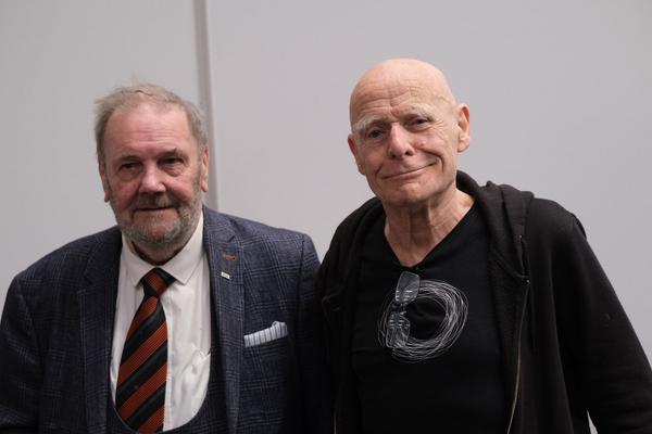 Jim and Eamonn pose for a photo, Jim wears a dark coloured suit, white shirt and striped tie, Eamonn wears dark coloured clothing. 