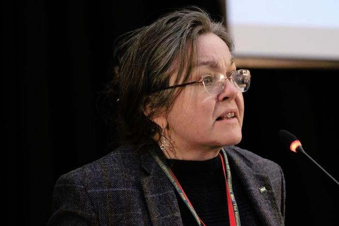 Pennie Quinton wearing a dark blazer and glasses speaks in opposition to motion two.