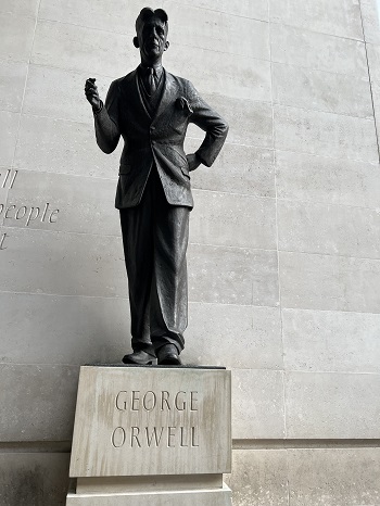 Statue of Orwell with one arm on hip and other raised. Orwell is depicted wearing a suit whilst standing on a stone square with his name inscribed.