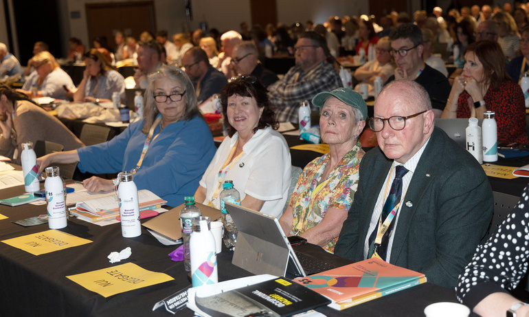 NUJ delegates sit at table with notepads and laptops as they pose for a photo. Others visible sitting at tables behind them. 