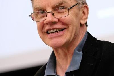 Simon smiles at podium. He wears a dark coloured shirt.