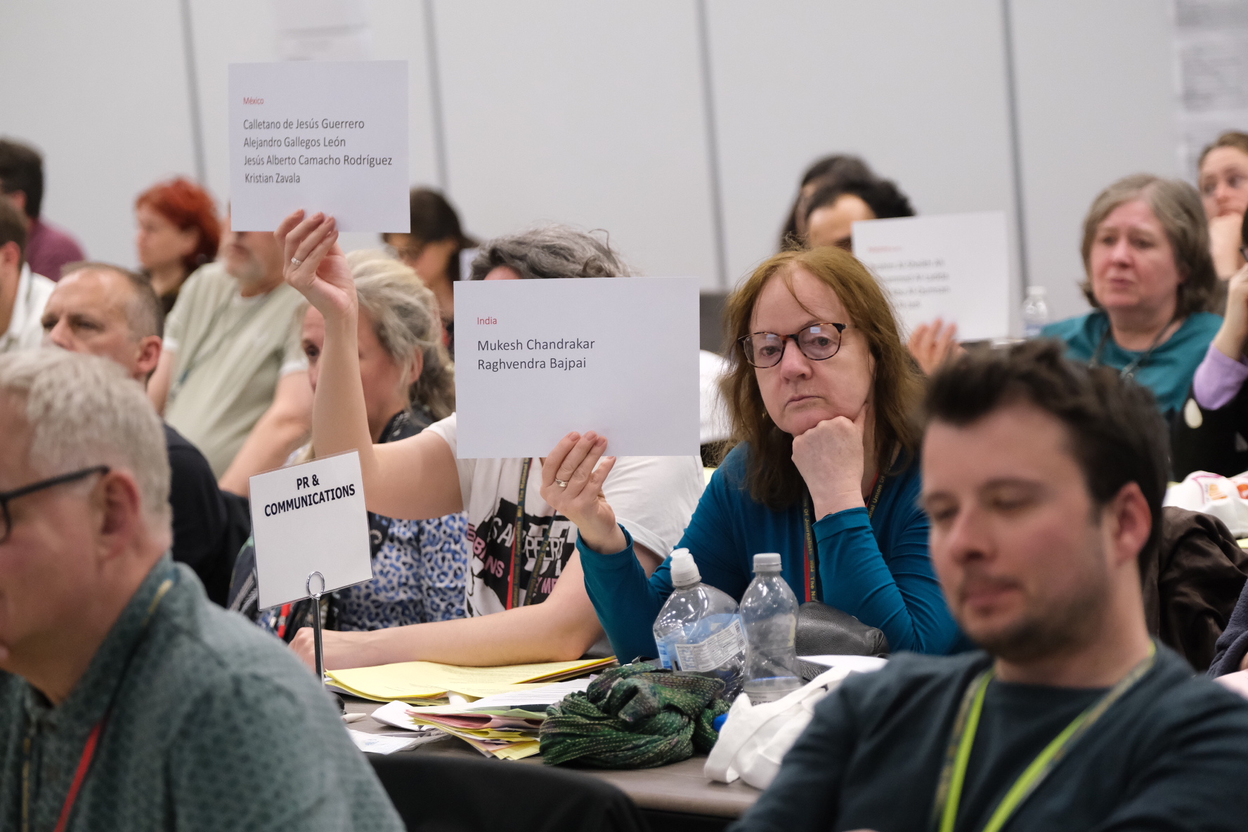 Delegates sit at tables whilst holding up landscape pieces of paper with the names of killed journalists.