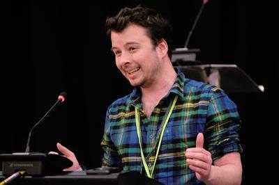 Sam wears a striped shirt and speaks at lectern with hands raised.