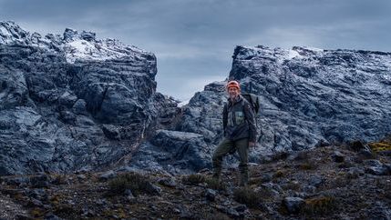 Klaus Thymann wearing hiking gear standing in front of mountainous, snow-topped rocks against a dark grey sky.
