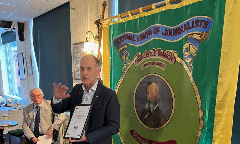 Jeff Brown wears dark suit and stands in front of green and yellow display whilst holding his life membership certificate,