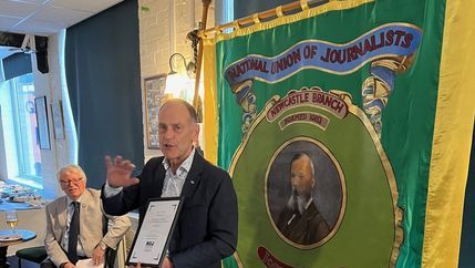 Jeff Brown wears dark suit and stands in front of green and yellow display whilst holding his life membership certificate,