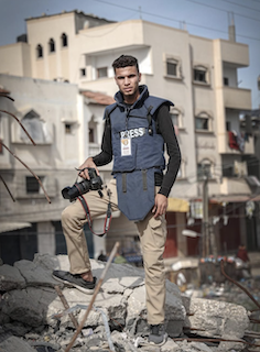 journalist Omar Al-Dirawi wears a blue PRESS vest whilst holding a black camera, standing on rubble. 