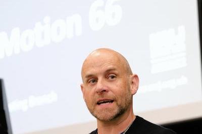 Trevor wears a dark jumper as he speaks into mic at the lectern.