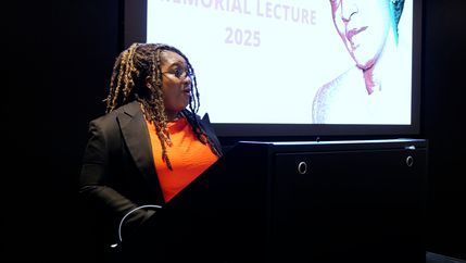 Bell Ribeiro-Addy MP wearing a dark blazer with an orange top stands in front of a podium.