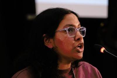 Saadeya wears maroon jumper and dark glasses as she speaks into black mic at lectern