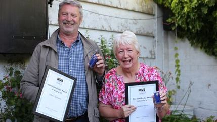Gilbert Johnston wearing a grey jacket and Caroline Sword wearing a pink top hold their NUJ gold badges and certificates.