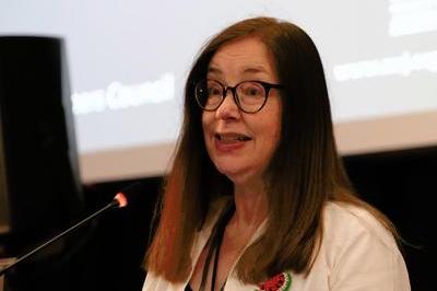 Ann wears a white blazer, has long shoulder length brown hair and wears dark coloured glasses as she presents at the lectern. Behind her 'motion 48' can be seen on the screen.