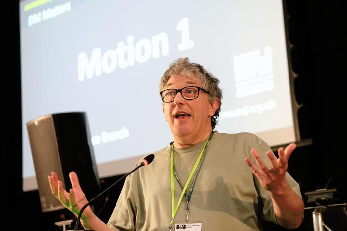 Mark Fisher wearing a grey t-shirt and glasses speaks at DM 2025 on behalf of the Edinburgh Freelance Branch
