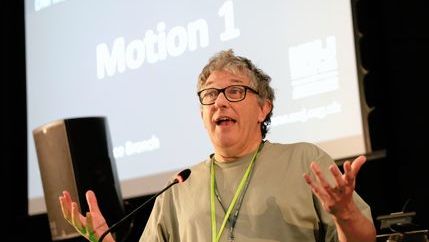 Mark Fisher wearing a grey t-shirt and glasses speaks at DM 2025 on behalf of the Edinburgh Freelance Branch