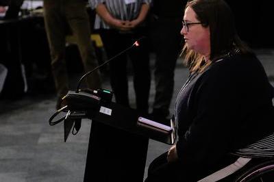 Gemma addresses delegates from her wheelchair. She speaks into mic at the lectern.