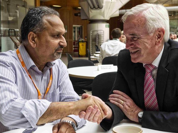 Barnie Choudhury and John McDonnell sitting at a table and shaking hands.
