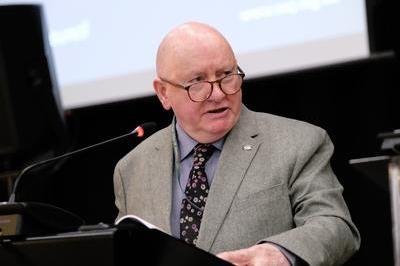 Séamus wears a grey suit and patterned tie, dark round glasses as he speaks at lectern.
