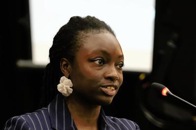 Cordu wears striped blazer and large floral earrings as she speaks into lectern.