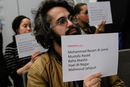 Man in beige jacket wearing dark glasses holds sign of the names of journalists killed. 