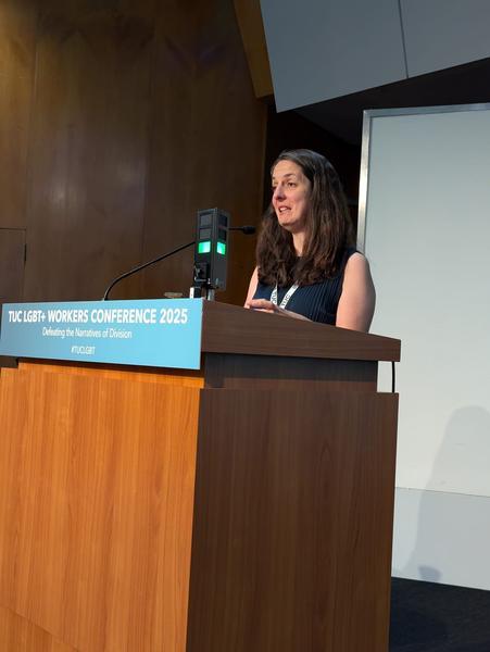 Natasha Hirst wearing a black top stands behind a podium at the TUC LGBT+ Conference 2025