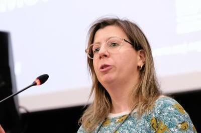 Cristina wears a patterned jumper and dark glasses as she speaks into a lectern. 