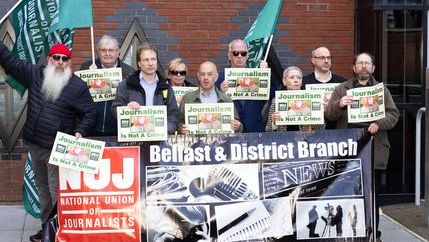 Attendees hold posters and wave NUJ flags as they pose for a photo with NUJ Belfast& District branch banner with red NUJ logo.