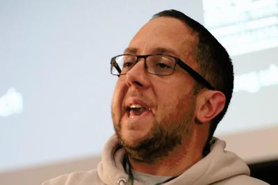 Matt wears a beige hoodie and holds notepad whilst speaking at the lectern.
