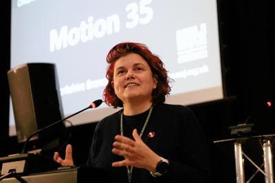 Katie speaks at lectern, she wears dark coloured jumper and has bright red hair.