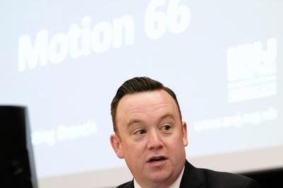 Fran wears a white shirt and dark suit with purple tie as he speaks into mic at the lectern.