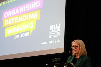 Rebecca presents from lectern, she wears a green blazer and dark glasses. Behind her is a screen with text organising, defending, winning and the NUJ logo.