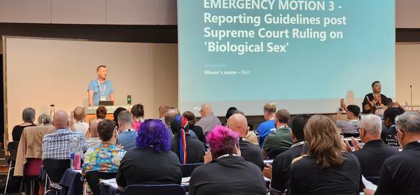 James Doherty in a blue shirt stands in front of a podium with an an audience in the foreground.
