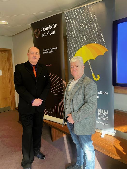 Gerry Curran wearing a black suit stands next to Gerard Cunningham wearing jeans and a grey blazer with an NUJ banner in the background.