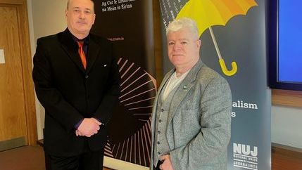 Gerry Curran wearing a black suit stands next to Gerard Cunningham wearing jeans and a grey blazer with an NUJ banner in the background.