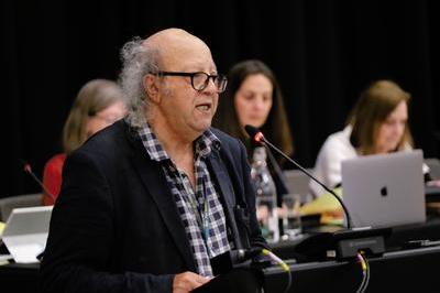 Jim Boumelha wears a dark blazer, checked shirt and dark glasses as he speaks at lectern