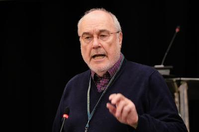 Chris wears dark jumper and black glasses as he speaks whilst pointing from the podium. 