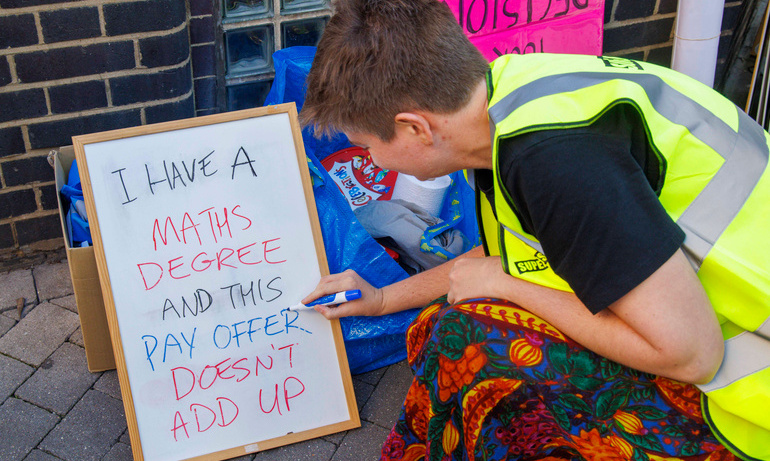 Journalist with brown short hair wearing red and black patterned dress writes on board ' I have a maths degree and this pay offer doesn't add up'