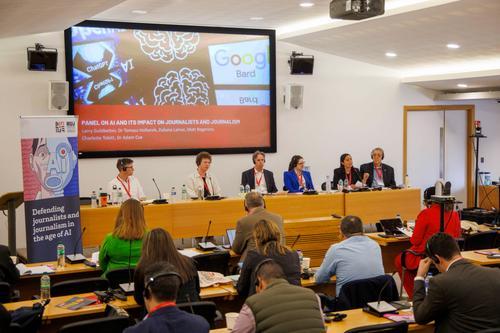 Audience face panellists sitting at desk with headsets. Behind them is a large slide with text panel on AI and its impact on journalists and journalism in white on red background.