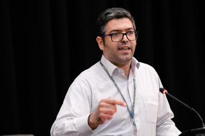 Behrang wears a white shirt and green lanyard dark glasses as he speaks into mic at lectern.